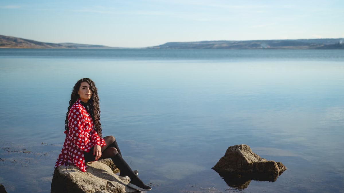 A woman in a red polka dot dress seated on rocks by a calm lake under a clear sky.
